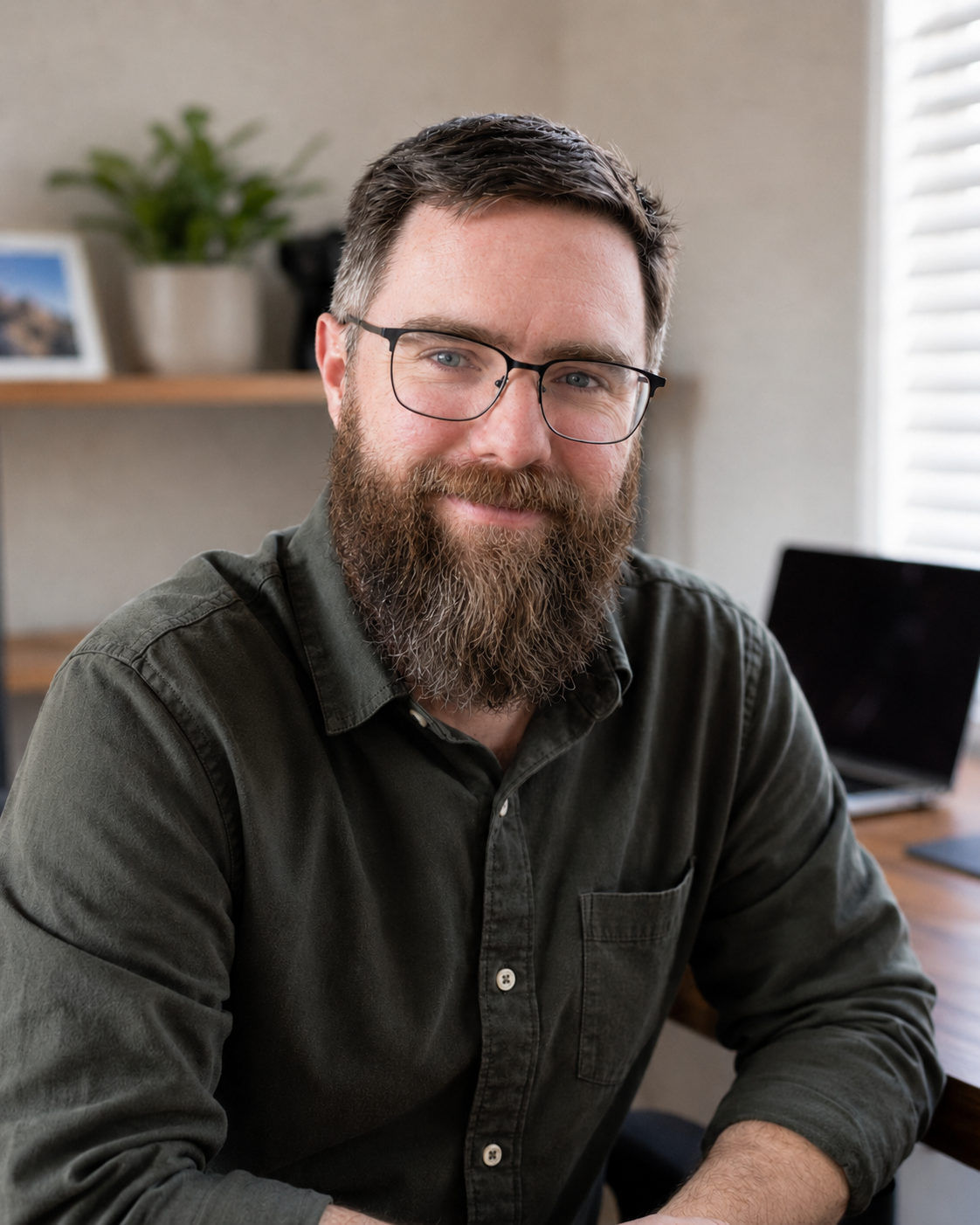John, a bearded man in glasses and an olive shirt, seated at his desk with a laptop behind him.